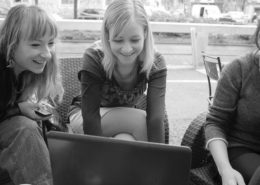 Drei junge Frauen sitzen am Nachmittag im Freien an einem Laptop und lachen gemeinsam, während sie auf den Bildschirm blicken.