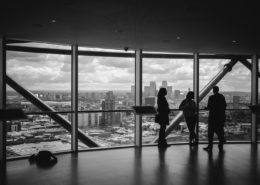 Drei Personen stehen am Nachmittag in einem hohen Gebäude vor einer großen Fensterfront und blicken auf die Skyline einer modernen Großstadt.