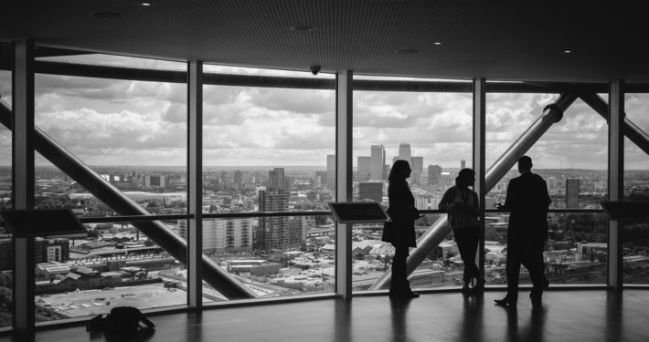 Drei Personen stehen am Nachmittag in einem hohen Gebäude vor einer großen Fensterfront und blicken auf die Skyline einer modernen Großstadt.