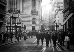Zahlreiche Menschen überqueren am späten Nachmittag eine belebte Straße im Stadtzentrum, während Sonnenlicht durch die Häuserschluchten fällt.