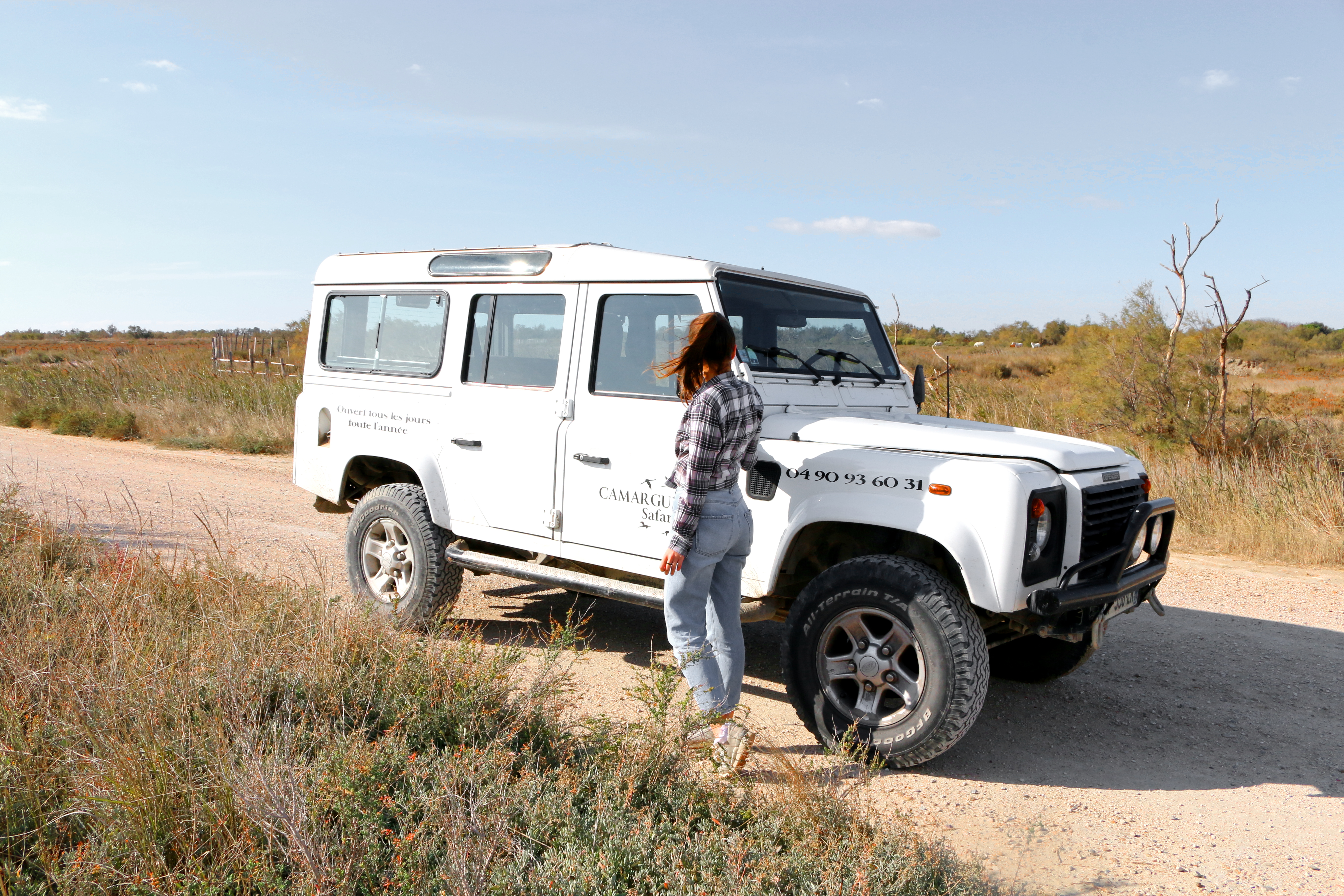 Eine Frau steht an einem weißen Jeep mit der Aufschrift „Camargue Safari“ auf einer staubigen Piste inmitten der Natur.