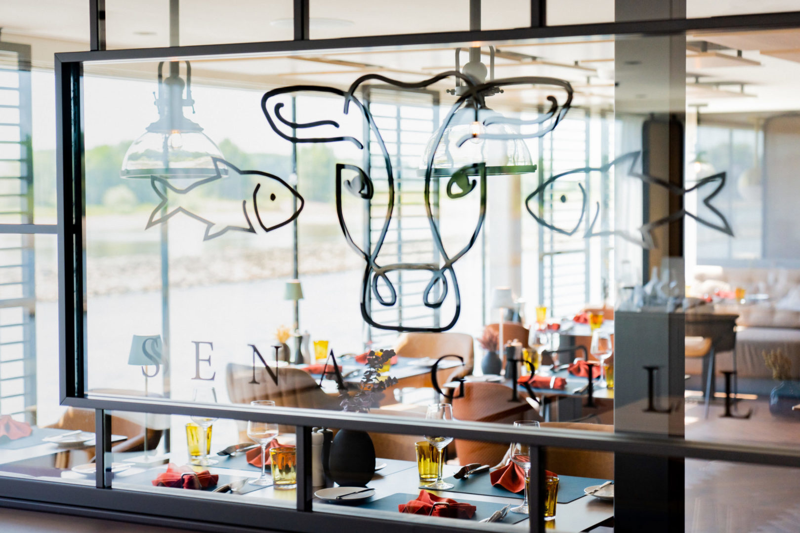 Blick durch eine Glaswand mit Tiermotiven ins stilvoll eingedeckte Restaurant SENA GRILL an Bord der A-ROSA SENA bei Tageslicht.