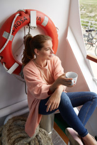 Frau sitzt auf einem Boot neben einem roten Rettungsring mit der Aufschrift 'Arosa' und hält eine weiße Tasse