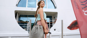 Frau mit langem Haar, trägt hellblaues Top und rosa Hose, steht vor einem weißen Schiff mit rundem Fenster, hält graue Handtasche und schwarzen Rollkoffer