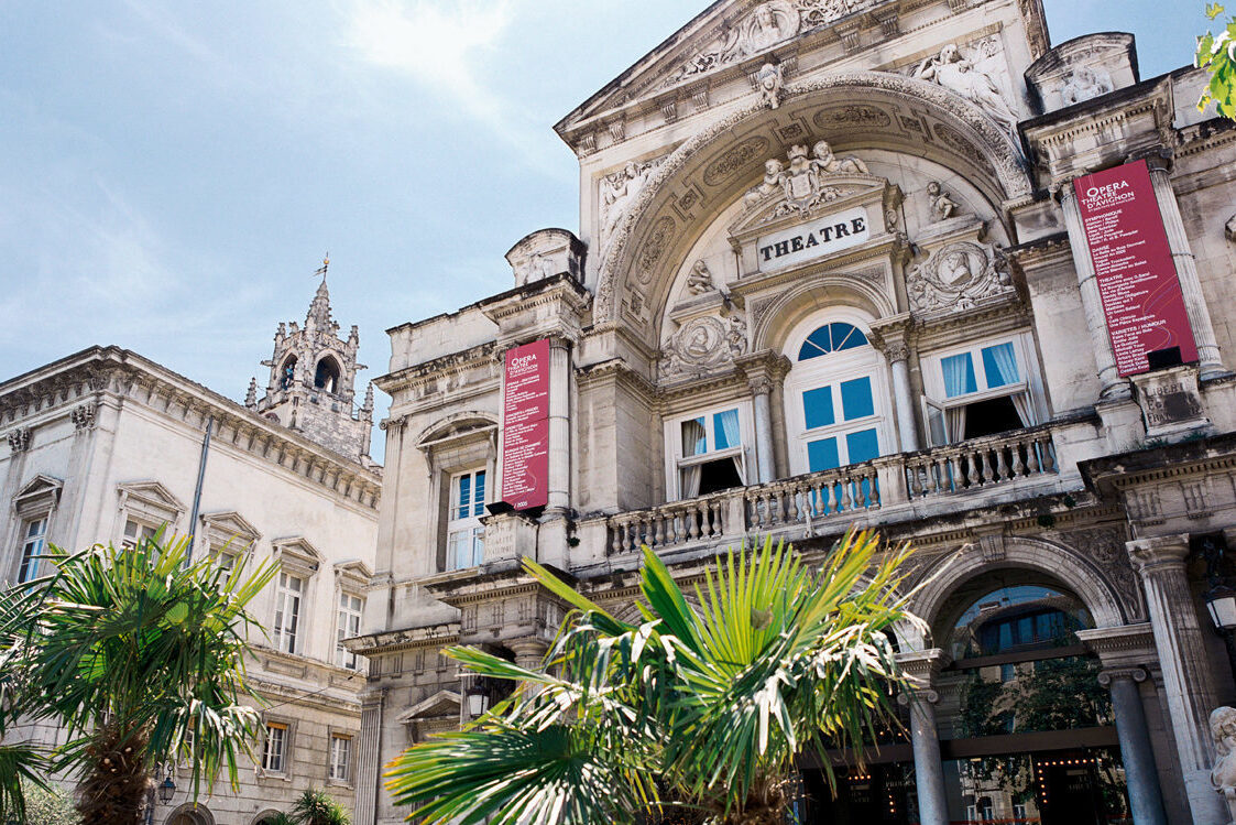 Die historische Oper von Avignon zeigt sich am späten Vormittag unter blauem Himmel, eingerahmt von Palmen und barocken Fassadendetails.