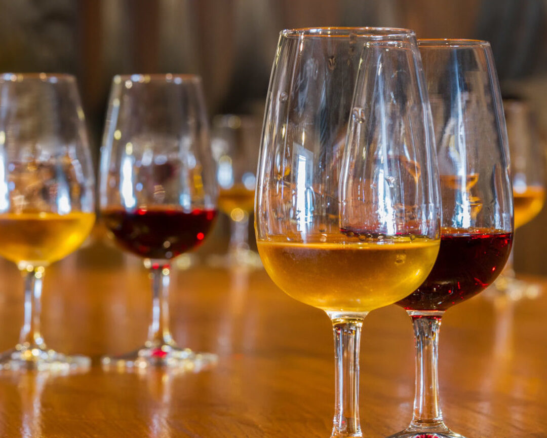 Mehrere Weingläser mit Portwein in verschiedenen Farben auf einem Holztisch, unscharfer Hintergrund mit Weinfässern