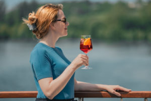 Frau mit Sonnenbrille und hochgestecktem Haar hält ein Glas mit rotem Getränk und Orangenscheibe am Geländer mit Wasser im Hintergrund