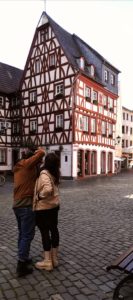 Zwei Personen betrachten ein historisches Fachwerkhaus mit roten Holzbalken und weißen Wänden an einem gepflasterten Platz