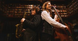 Zwei Musiker stehen Rücken an Rücken, einer spielt Saxophon, der andere Kontrabass, vor einer Wand voller Bücher