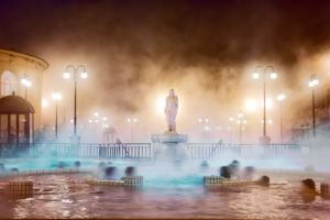 Dampfende Thermalquelle mit Menschen im Wasser und einer Statue in der Mitte, umgeben von historischen Gebäuden und Straßenlaternen bei Nacht