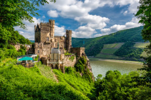 Burg Rheinstein auf einem bewachsenen Felsen mit Blick auf den Rhein und bewaldete Hügel unter blauem Himmel mit Wolken