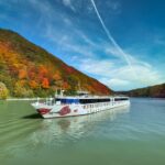 Flusskreuzfahrtschiff auf ruhigem Fluss vor herbstlich bunt gefärbtem Wald und blauem Himmel mit Wolken