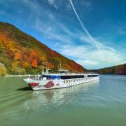 Flusskreuzfahrtschiff auf ruhigem Fluss vor herbstlich bunt gefärbtem Wald und blauem Himmel mit Wolken