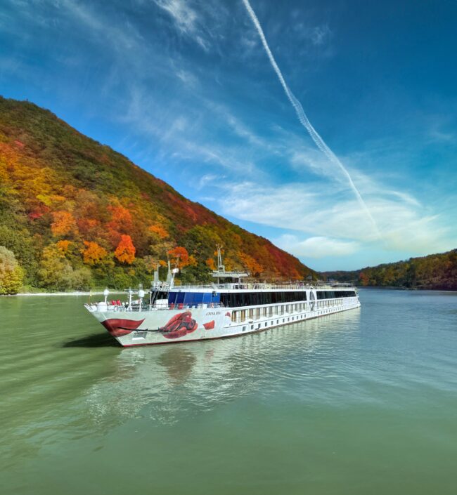 Flusskreuzfahrtschiff auf ruhigem Fluss vor herbstlich bunt gefärbtem Wald und blauem Himmel mit Wolken
