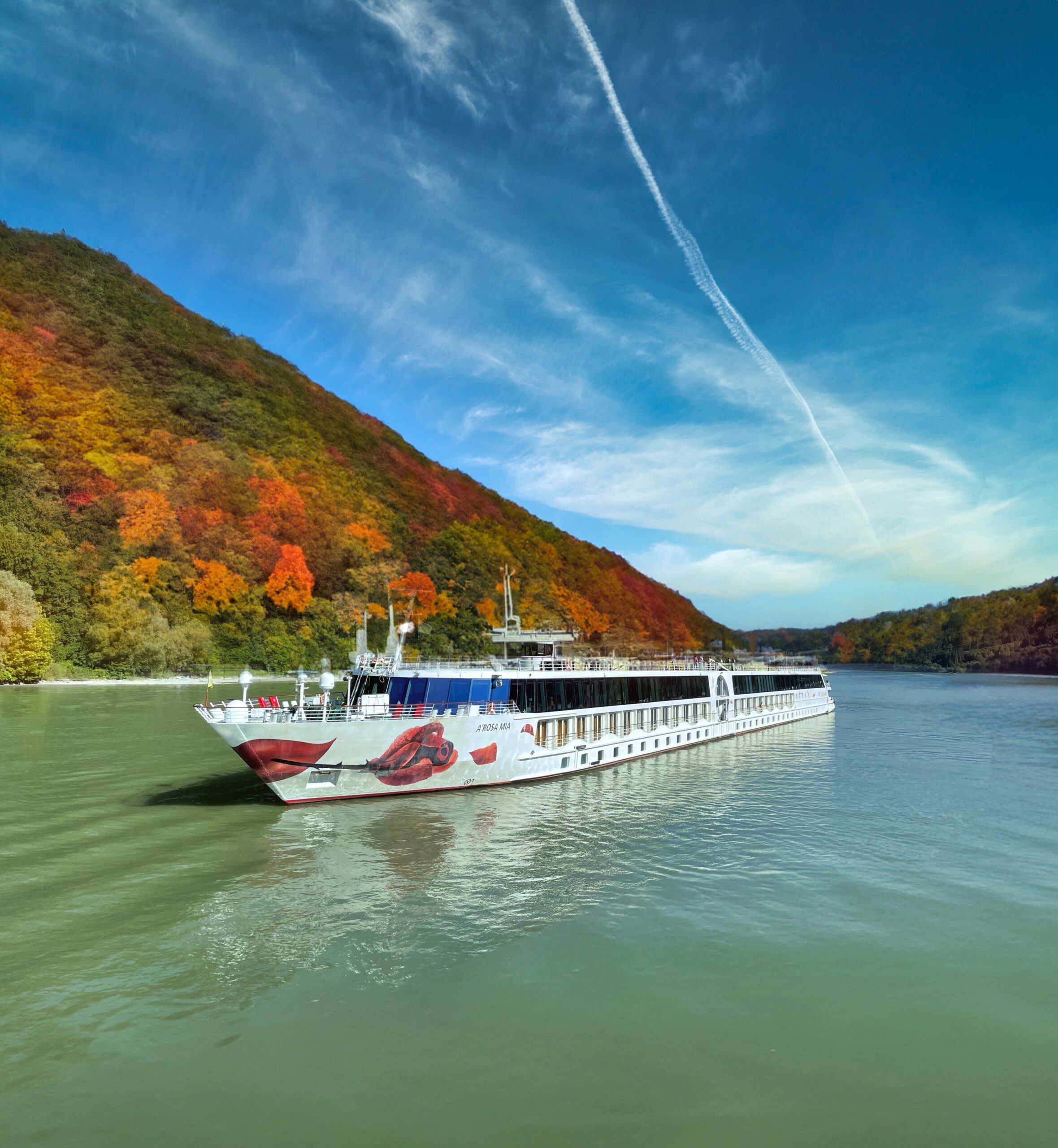 A-ROSA MIA im Herbst Flusskreuzfahrtschiff auf ruhigem Fluss vor herbstlich bunt gefärbtem Wald und blauem Himmel mit Wolken