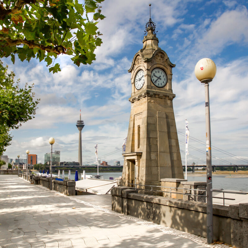 Pegeluhr am Rhein in Düsseldorf Die historische Pegeluhr an der Rheinuferpromenade in Düsseldorf zeigt am späten Vormittag die Zeit, im Hintergrund sind Rheinturm und Brücke sichtbar.