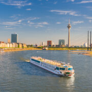 Ein A-ROSA Schiff fährt am späten Nachmittag über den Rhein in Düsseldorf, im Hintergrund sind der Rheinturm und die Stadtsilhouette zu sehen.