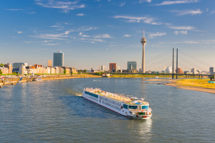 Ein A-ROSA Schiff fährt am späten Nachmittag über den Rhein in Düsseldorf, im Hintergrund sind der Rheinturm und die Stadtsilhouette zu sehen.