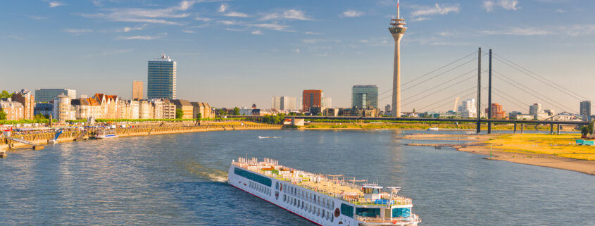 Ein A-ROSA Schiff fährt am späten Nachmittag über den Rhein in Düsseldorf, im Hintergrund sind der Rheinturm und die Stadtsilhouette zu sehen.