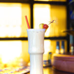 Ein tropischer Cocktail (Charlie Brown) mit Ananas und Kirsche wird am späten Nachmittag vor leuchtend gelbem Fensterlicht auf einer Bar serviert.