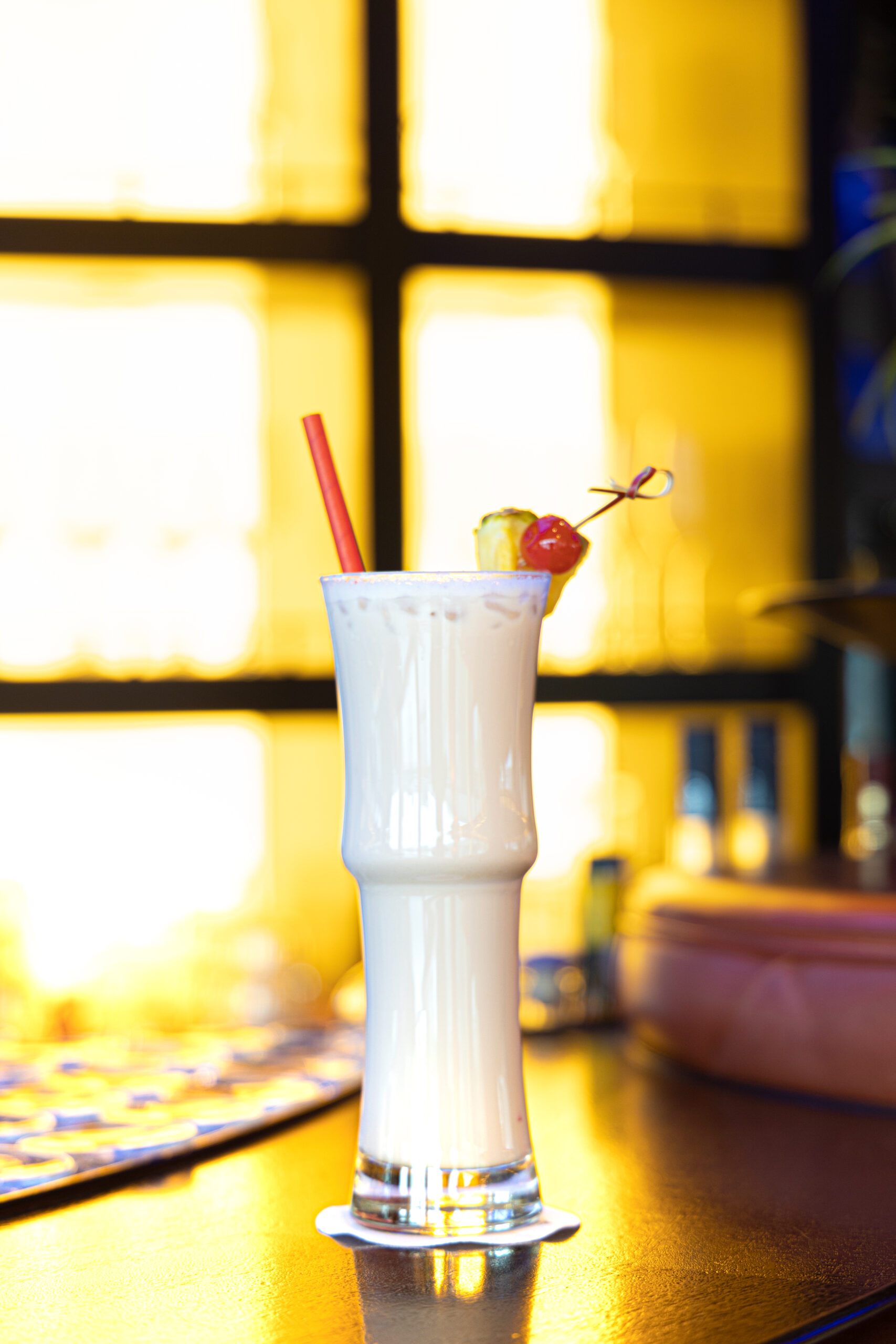 Charlie Brown Ein tropischer Cocktail (Charlie Brown) mit Ananas und Kirsche wird am späten Nachmittag vor leuchtend gelbem Fensterlicht auf einer Bar serviert.