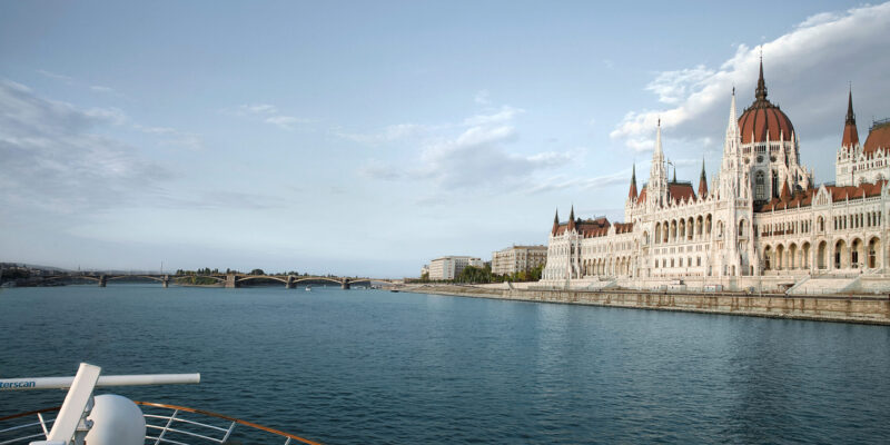 Blick von einem Kreuzfahrtschiff auf das Parlamentsgebäude in Budapest am Donauufer unter bewölktem Himmel.