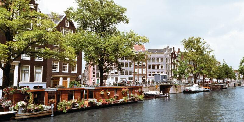Ansicht von einer Gracht in Amsterdam mit traditionellen Häusern und Hausbooten bei bewölktem Himmel. Am Ufer sind Bäume, Fahrräder und Passanten.